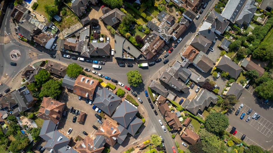 An aerial view of a residential area showing tightly packed houses with pitched roofs, some with chimneys, surrounded by trees and gardens. A narrow, winding street runs through the neighborhood, with several parked cars lining the road and in designated parking areas. The street appears to be in the process of a home relocation, with a series of moving vehicles, including a white van, visible along the roadway. In the foreground, a small parking lot contains additional vehicles, and the surrounding properties feature various materials such as brick, slate, and tile roofs, with some houses having visible driveways and front gardens. The scene captures the typical infrastructure of an urban suburb, with clean, well-maintained streets, and lush greenery. This setting is relevant to house removals and furniture transport services provided by companies like Man with Van Chislehurst, offering an overview of the area where local moving and packing operations are often conducted.