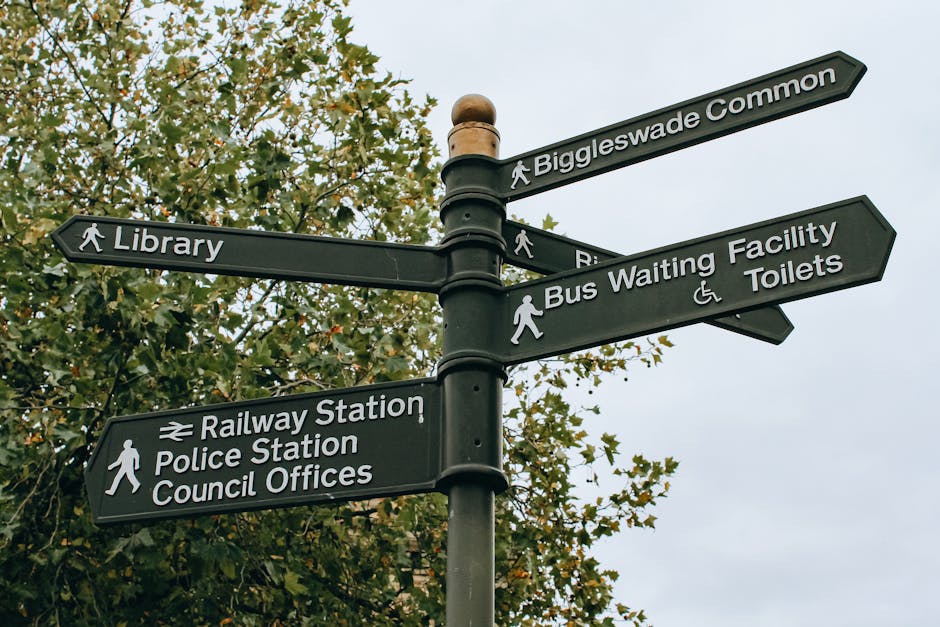 A black multi-directional signpost with white text indicating various local destinations including Biggleswade Common, Library, Bus Waiting Facility and Toilets, Railway Station, Police Station, and Council Offices, each with associated icons for pedestrians or facilities. The signpost is situated outdoors against a backdrop of leafy trees with green foliage, under a cloudy sky. The signs are mounted on a tall pole and point in different directions, providing guidance for pedestrians and visitors. This image relates to house removals and relocation services operated by Man with Van Chislehurst, illustrating local area navigation relevant to moving logistics and home relocation processes within the community.