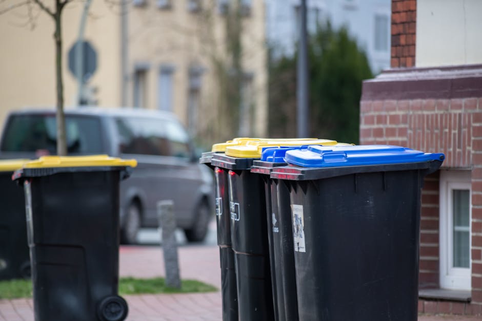 A row of three large wheeled waste bins with black bodies and yellow, blue, and black lids positioned outdoors on a residential pavement. The bins are lined up close to the edge of a brick building with part of the brick wall visible on the right. Behind the bins, a grey van is parked on the street, slightly out of focus. In the background, there are other residential buildings, trees, and a sidewalk. The scene is lit with natural daylight, indicating daytime. This image captures waste disposal in a suburban area, relevant to house removals and the disposal of bulky waste or mattresses as outlined in Chislehurst's waste disposal rules. Man with Van Chislehurst occasionally assists with home relocation, including loading and transport processes.