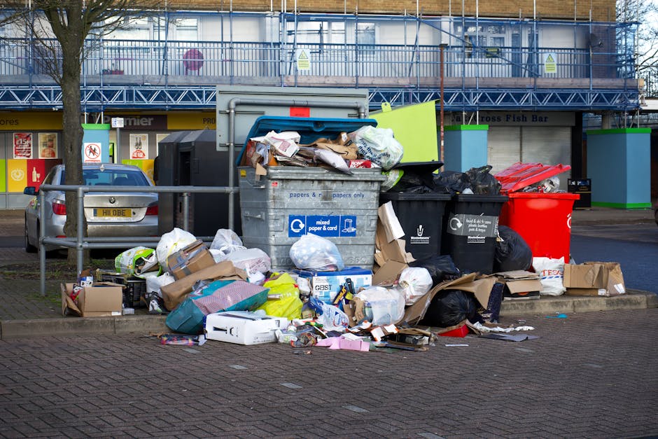 An outdoor scene showing overflowing waste and recycling bins situated on a paved area beside a sidewalk in Chislehurst. The bins include a large grey mixed paper and cardboard container, a black recycling bin, a bright red waste bin, and a black bin with a flap lid, all of which are overfilled with paper, cardboard, plastic bags, and packaging materials. Cardboard boxes and loose packaging are scattered on the ground in front of the bins, along with crumpled paper, plastic bottles, and other miscellaneous rubbish. Behind the waste area, a parked silver car is visible, with a protective metal railing separating it from the spill. The background features a commercial building with signage and a construction scaffold covering part of its facade. The scene reflects typical household or business waste disposal practices, relevant to house removal or furniture transport preparations, as managed by companies like Man with Van Chislehurst.
