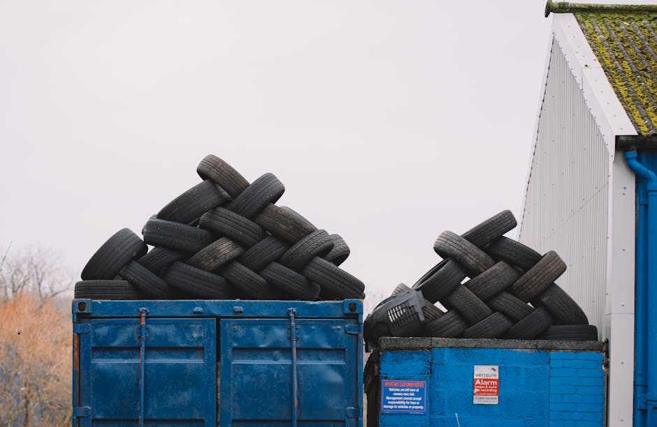 A row of three large wheeled waste bins with black bodies and yellow, blue, and black lids positioned outdoors on a residential pavement. The bins are lined up close to the edge of a brick building with part of the brick wall visible on the right. Behind the bins, a grey van is parked on the street, slightly out of focus. In the background, there are other residential buildings, trees, and a sidewalk. The scene is lit with natural daylight, indicating daytime. This image captures waste disposal in a suburban area, relevant to house removals and the disposal of bulky waste or mattresses as outlined in Chislehurst's waste disposal rules. Man with Van Chislehurst occasionally assists with home relocation, including loading and transport processes.