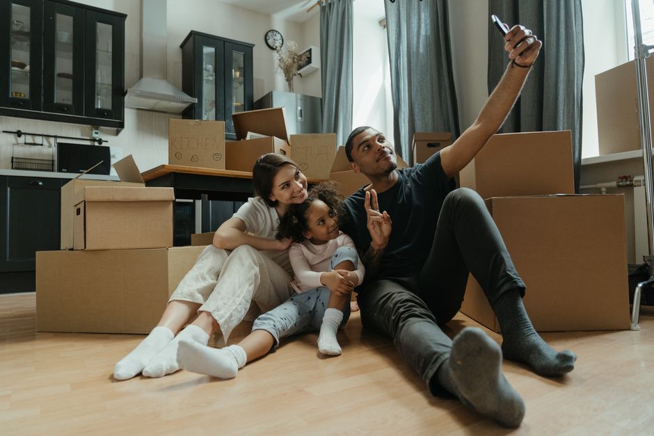 A young family sitting on a light wooden floor inside a well-lit kitchen with dark cabinetry and a window with blue curtains. The family consists of a mother, father, and a young girl, all smiling and relaxed. The father, wearing a black t-shirt and grey socks, is holding a smartphone in his extended right hand, taking a selfie while making a peace sign with his left hand. The mother, dressed in a white top and light-colored pants, is leaning close to her daughter, who is sitting between them in light pajamas, with curly hair, smiling and looking up at the father. Surrounding them are several large cardboard boxes and packing materials, indicating a home relocation or move-in process, with some boxes stacked and others open. The scene captures a moment of packing, unpacking, or documenting the moving process as part of professional house removal services by Man with Van Chislehurst, in a neutral and natural manner.