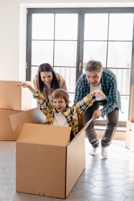 A family of three, including a young boy in a yellow checkered shirt sitting inside an open cardboard moving box, with arms raised in excitement. Behind them, a woman with long dark hair and a man with short gray hair, both smiling, are assisting with packing or home relocation inside a well-lit room with large glass doors. The room features a tiled floor and is filled with several other packed cardboard boxes of various sizes, indicating ongoing packing or furniture transport preparations. The scene reflects a positive atmosphere of packing and moving activities associated with house removals, with the family engaged in the process of a home relocation supported by professional moving services such as those offered by Man with Van Chislehurst.
