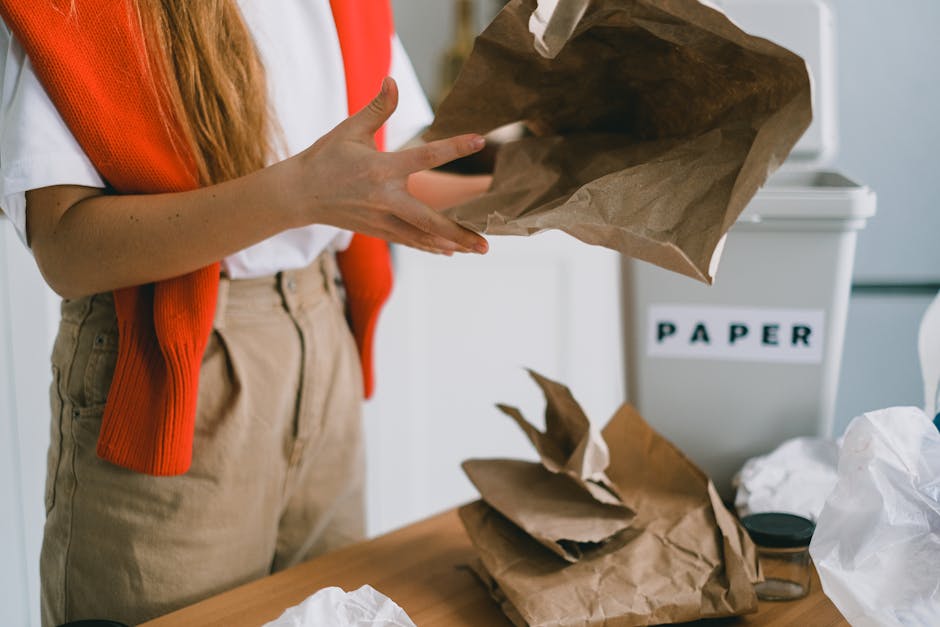 A person wearing a white shirt and beige trousers, with an orange cardigan draped over their shoulders, is placing a crumpled brown paper bag onto a table inside a property. On the table, there are ad