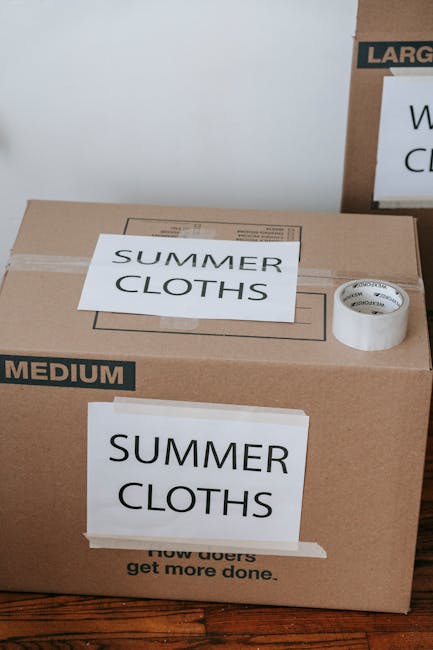 A cardboard box labeled 'SUMMER CLOTHS' and 'SUMMER' is placed on a wooden floor inside a property, with a roll of packing tape and a smaller cardboard box on top of the larger box. The packaging materials are white with black text, and the larger box is marked 'MEDIUM' in black on the side. In the background, there is another cardboard box partially visible with additional packing labels. This scene depicts the packing stage of a home relocation managed by Man with Van Chislehurst, with boxes ready for moving. The lighting is natural, and the area appears organized for a furniture transport or packing and moving process, as part of a house removal or moving service, with focus on the careful packing of clothes for transport.