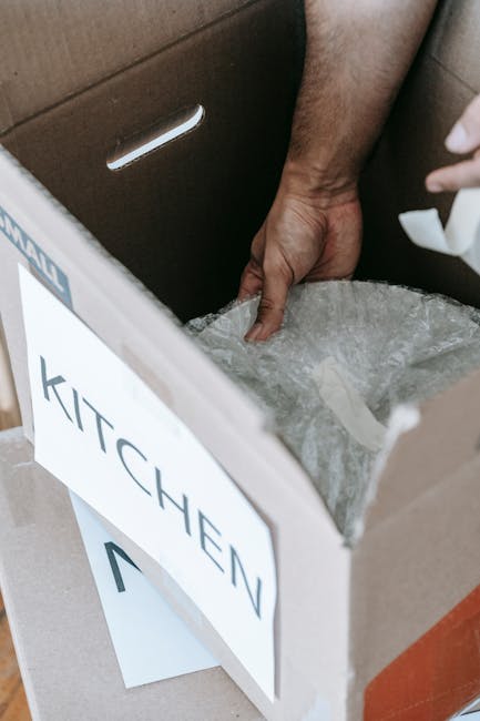 Close-up of a person's hand placing a cardboard box labeled 'KITCHEN' onto a surface inside a property, with the box partially open revealing packing material such as bubble wrap inside. The box is oriented with the label facing outward for easy identification during the home relocation process. In the background, part of a brown cardboard moving box and a segment of a door frame or wall is visible, indicating the interior of a house being prepared for moving. The scene captures the packing phase of furniture transport, involving careful handling of items and materials, and is associated with professional removals services like those provided by Man with Van Chislehurst. The image emphasizes organized packing and secure loading in the context of house removals in Chislehurst.
