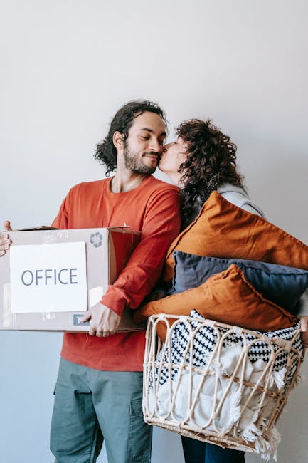 A man with a beard, wearing a red long-sleeve shirt and gray pants, is holding a cardboard box labeled 'OFFICE' in black text, with the recycling symbol on it. A woman with curly dark hair, dressed in a gray top and blue jeans, is leaning in to kiss him on the cheek. She is carrying a large woven basket filled with various cushions and folded blankets, some of which are orange, navy blue, and striped. They are standing indoors against a plain white wall, and the scene captures a moment of packing or home relocation preparation, reflecting furniture transport and moving logistics in a professional house removal context. The image naturally supports themes of packing, moving, and household logistics, as would be relevant for a company like Man with Van Chislehurst working within removals services.