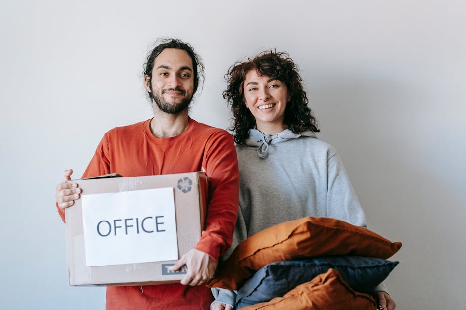 A man and a woman standing indoors against a plain light grey wall, preparing for an office relocation. The man, with dark, curly hair and a beard, wears a red long-sleeve shirt and holds a cardboard box labeled 'OFFICE' with a recycling symbol in the top right corner. The woman, with short, curly brown hair, is dressed in a grey hoodie and holds several cushions wrapped in fabric, including orange and blue covers. Behind them, the area appears to be a room with ample natural lighting, and the focus is on their expressions and the packing materials, illustrating the process of furniture transport and packing during a professional move. This image reflects aspects of house and office removals, highlighting packing, loading, and moving logistics, with Man with Van Chislehurst's services supporting minimal downtime for SME relocations.