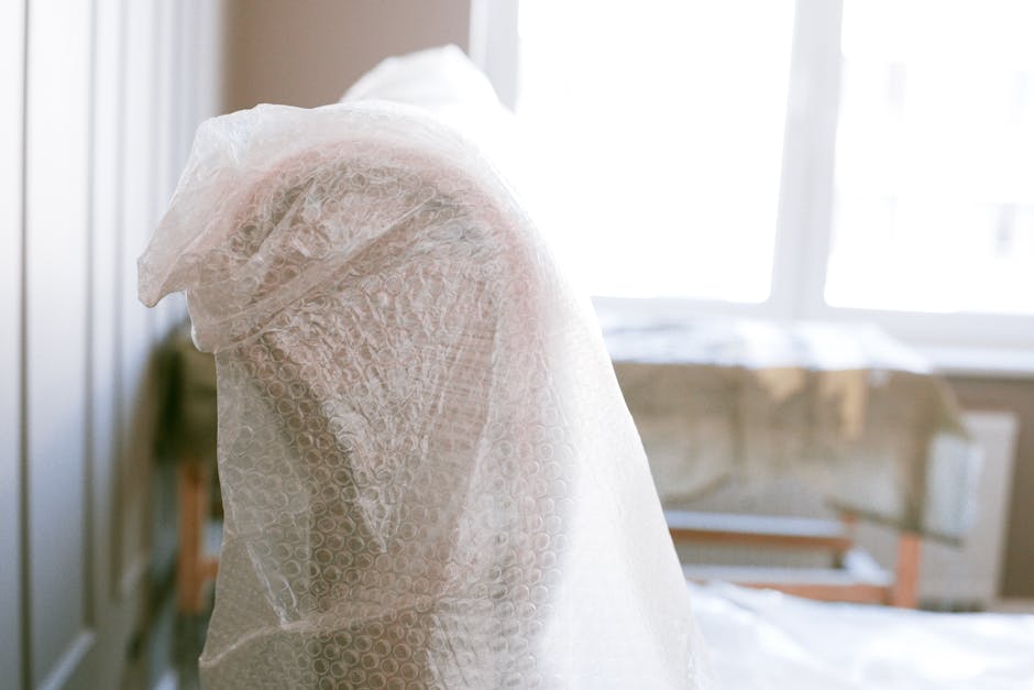A close-up of a piece of furniture wrapped securely in transparent bubble wrap, standing upright inside a well-lit room with natural daylight coming through large windows in the background. The bubble wrap covers the entire object, ensuring protection during the packing process. Nearby, partially visible, are additional packing materials such as cardboard boxes and fabric covers placed on the floor, indicating a home relocation or moving preparation. The environment appears calm and organized, with the furniture ready for transport, reflecting professional packing services provided by Man with Van Chislehurst as part of a house removals and furniture transport operation.