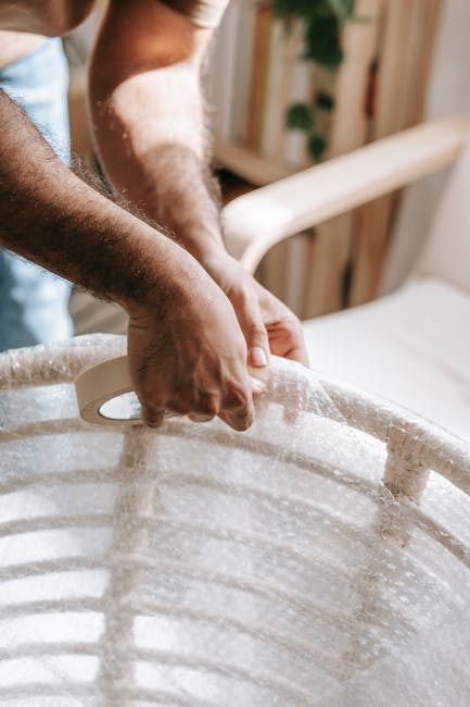Close-up of a person’s hands lifting a large, oval-shaped, light-colored cardboard container with reinforced edges, wrapped in protective bubble wrap, inside a room with wooden furniture and shelves in the background. The individual is handling the container carefully as part of a home relocation or furniture transport process, with a focus on packing fragile items such as antiques. The scene suggests the loading process during a house removal service, with natural lighting illuminating the workspace. In the context of house removals, this image illustrates careful packing and handling of delicate items before transport, aligning with professional packing and moving activities provided by Man with Van Chislehurst.