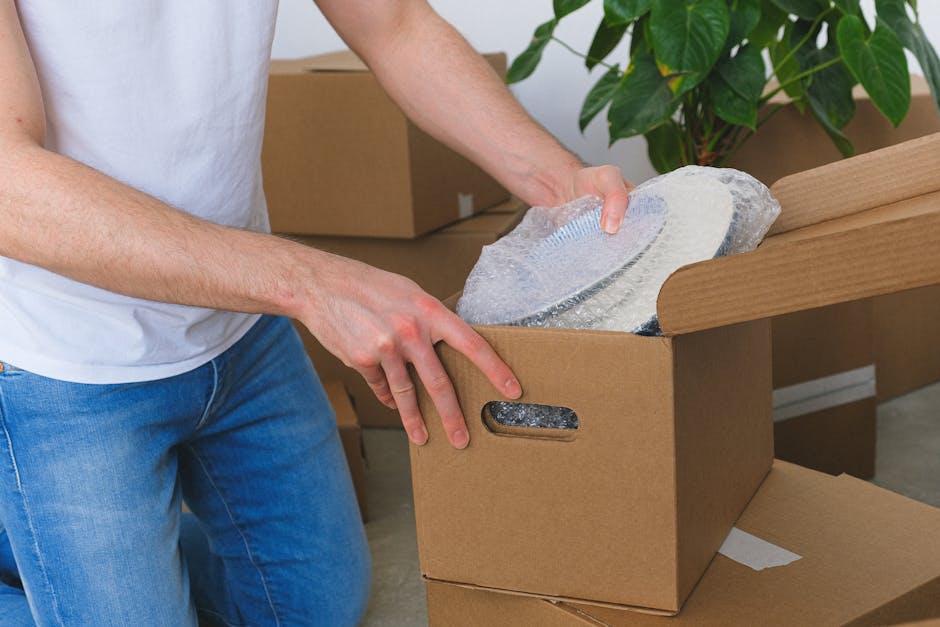 A close-up of a piece of furniture wrapped securely in transparent bubble wrap, standing upright inside a well-lit room with natural daylight coming through large windows in the background. The bubble wrap covers the entire object, ensuring protection during the packing process. Nearby, partially visible, are additional packing materials such as cardboard boxes and fabric covers placed on the floor, indicating a home relocation or moving preparation. The environment appears calm and organized, with the furniture ready for transport, reflecting professional packing services provided by Man with Van Chislehurst as part of a house removals and furniture transport operation.