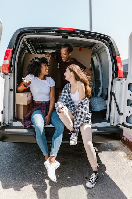 Three individuals, two women and one man, sit inside the open cargo area of a large grey van parked outside on a paved surface, with the van's interior filled with cardboard boxes and packing material