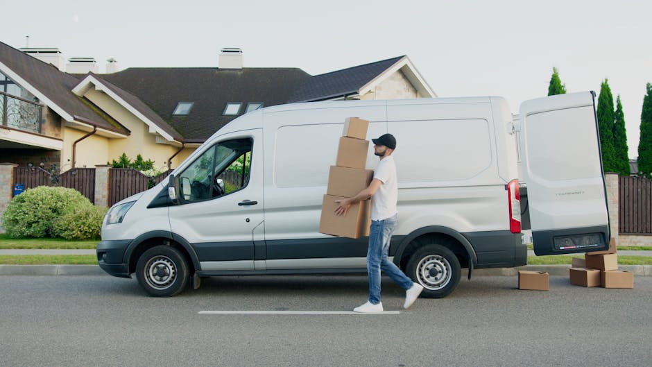 Three individuals, two women and one man, sit inside the open cargo area of a large grey van parked outside on a paved surface, with the van's interior filled with cardboard boxes and packing material