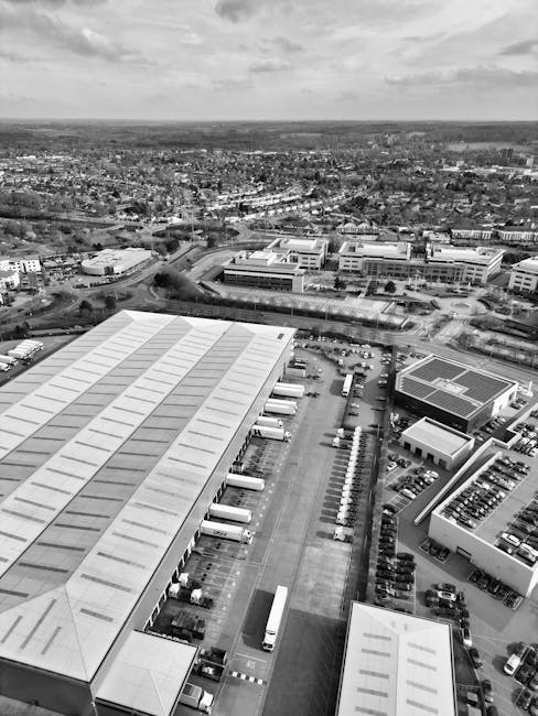 A black and white aerial view showing a large industrial warehouse with a long, pitched roof and multiple loading bays along its side. Several trucks are parked adjacent to the loading bays, some posi