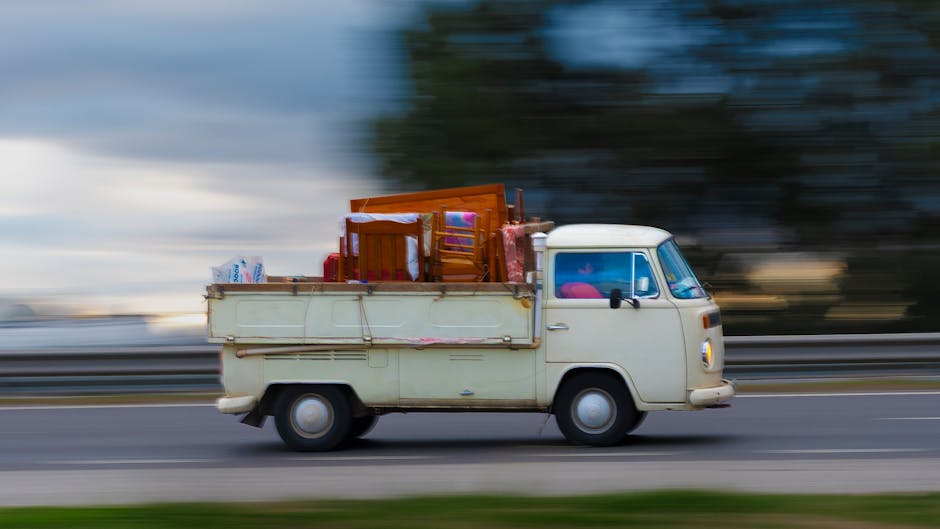 A vintage cream-coloured van with an open cargo area moving along a road, loaded with various household furniture including wooden chairs, a table, and household items wrapped in blankets and plastic covering. The van is in motion, indicated by the blurred background of trees and sky, and is positioned on a paved surface with metal guardrails visible behind. Inside the cargo area, the furniture is stacked and secured, typical of home relocation or furniture transport during packing and moving processes. The vehicle appears to be part of a professional removals service, such as Man with Van Chislehurst, involved in house removals or moving logistics, as seen on the webpage titled 'Why Licensed, Insured Removers Matter in Chislehurst.'