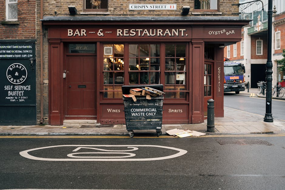 An exterior view of a traditional pub with a dark red wooden façade, large glass windows, and wooden framing, situated on a city street. A black waste bin labeled 'Commercial Waste Only' is placed on the pavement in front of the establishment, with cardboard boxes and paper packaging resting on top of it. To the left, there is a blackboard sign advertising a self-service buffet, and to the right, a black bollard and a lamppost are positioned near the sidewalk. A white road marking indicating a bus stop or no-parking zone is visible on the tarmac, and in the background, a delivery truck and parked bicycles can be seen along the roadside. The scene is lit by natural daylight, capturing a moment where items are temporarily outside the building, potentially during a home relocation or moving process managed by professional removers such as Man with Van Chislehurst.